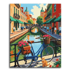 A paint by numbers canvas of a blue bicycle with pink flowers leaning on a canal railing, a ginger cat beside it, and a sunny European street with green awnings behind.