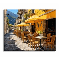 A cobbled street lined with bentwood chairs, tables and yellow umbrellas runs past a café toward a green mountain in the distance.